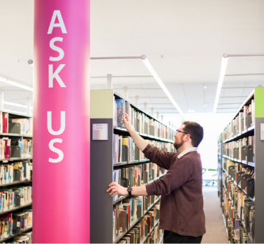 A man stands in a library infront of a sign that says Ask Us