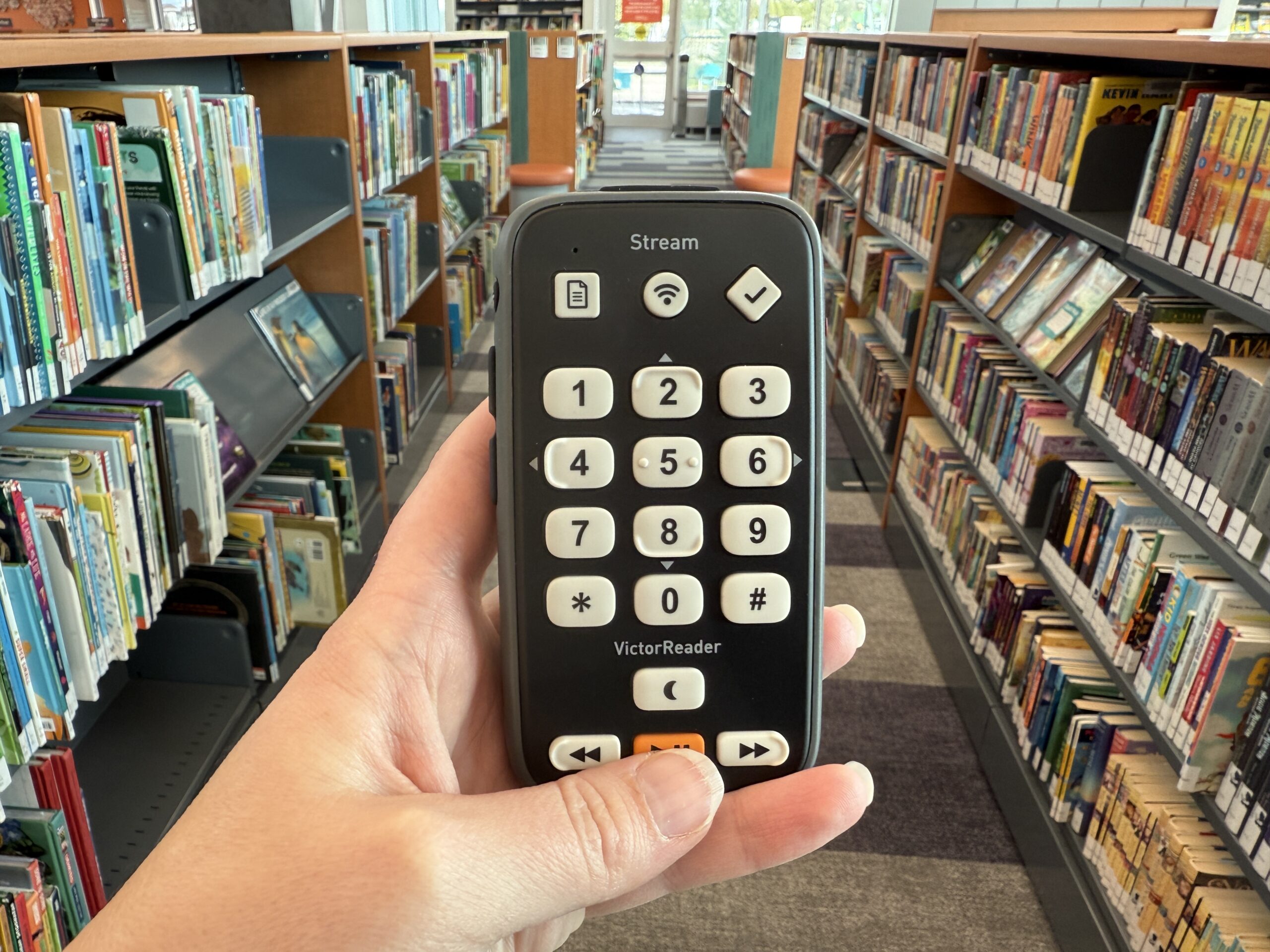 Close up of hand holding a Victor Reader Stream device in front of shelves of books.